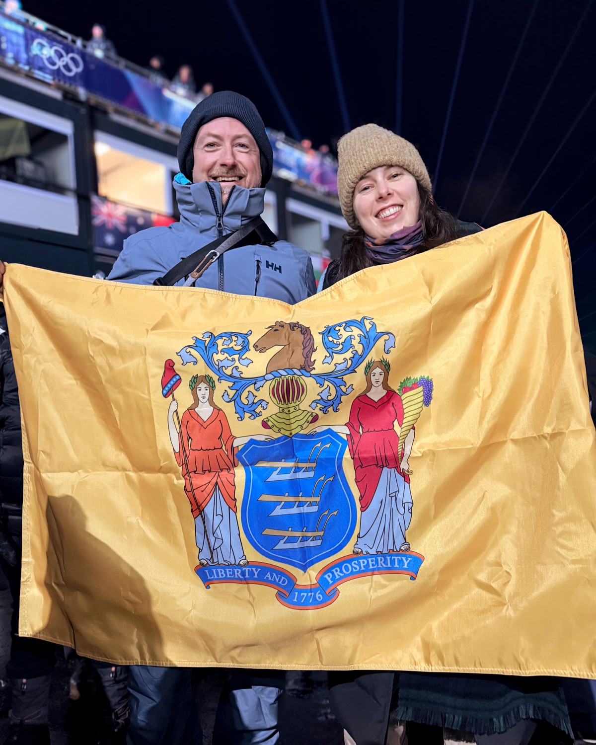 Richard and his wife at the 2026 Winter Olympics, holding the New Jersey state flag