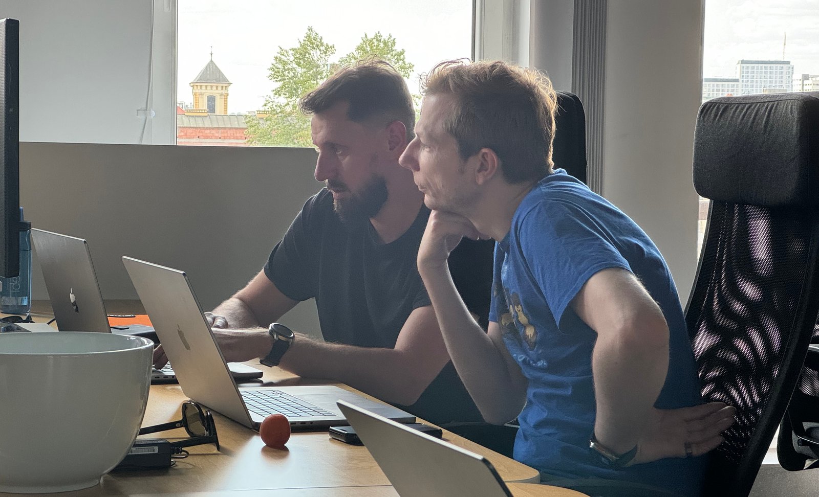 Markus and Michał focused on a shared laptop screen at Nomtek, Wrocław rooftops through the window behind them