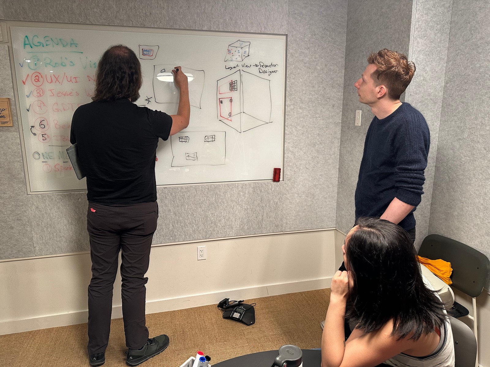 Three Slyyd team members at a whiteboard during an in-person workshop — one sketching an agenda and a lighting-setup diagram while two others watch